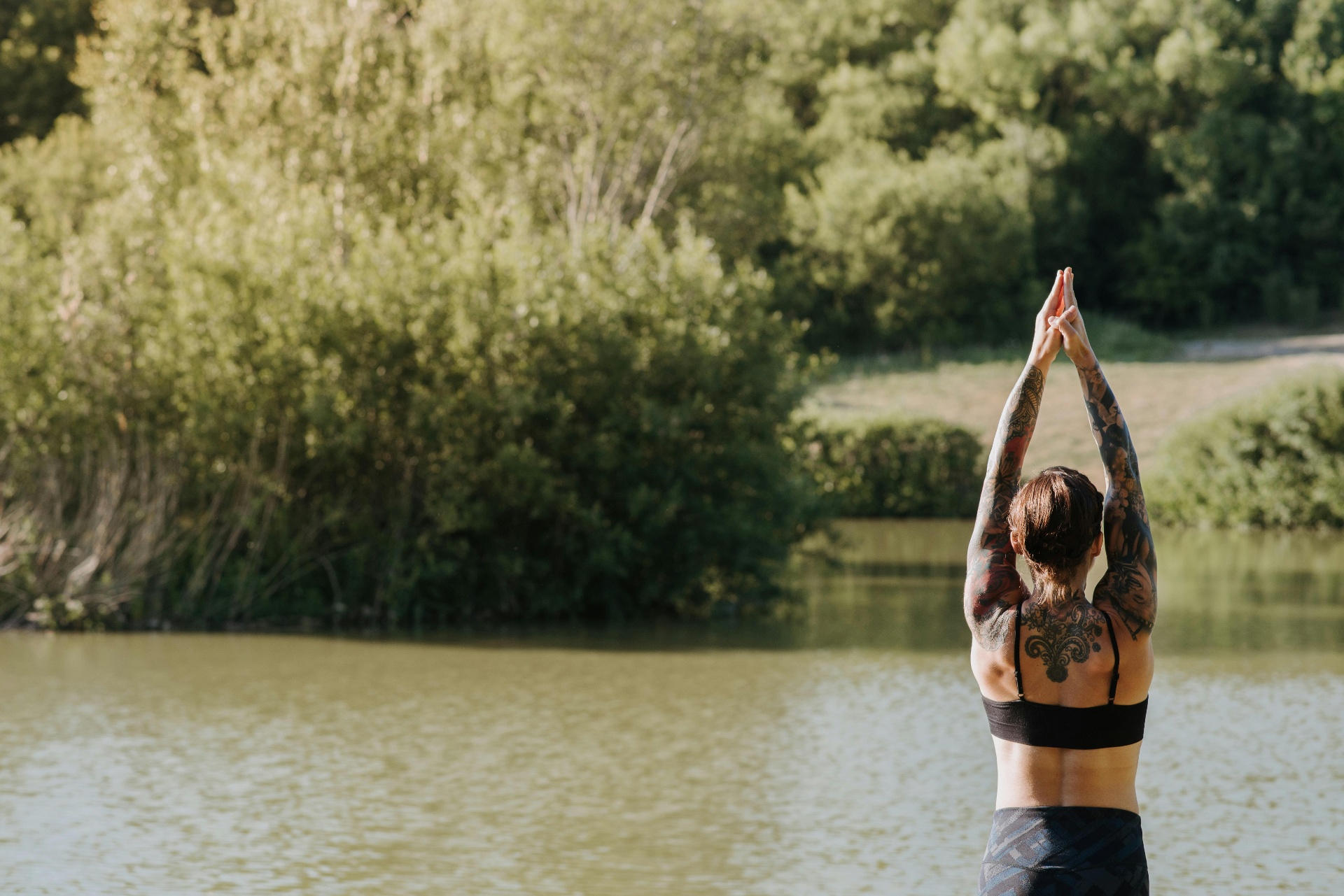 A tattooed woman in a black sports bra raising her arms overhead in a sun salutation by a tree-lined lake at golden hour